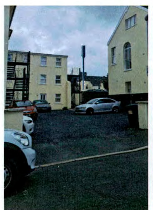 A photograph showing a paved parking area with several cars, flanked by multi-story buildings, with a tall telecommunication pole visible in the background.