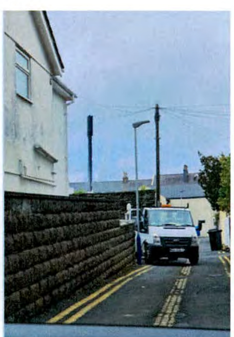 A street-level photograph showing a white building on the left adjacent to a dark retaining wall, with a white van on the road and a tall telecommunication pole visible in the background.