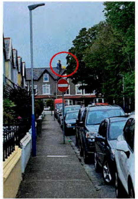 A street-level photograph showing a residential road with parked cars and terraced houses, featuring a red circle annotation on a distant roof.