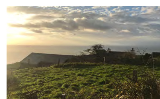 A photograph of a rural coastal landscape featuring a long stone building, possibly a barn, overlooking the sea at sunset.