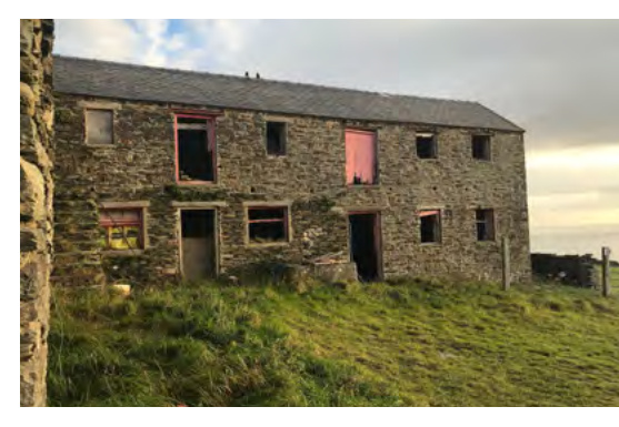 A photograph showing the exterior of a two-story stone building, likely a barn, situated on a grassy slope with open window frames.