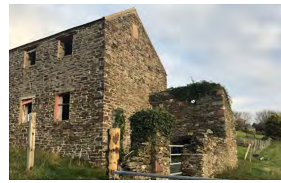 A photograph showing an existing two-story stone building with empty window openings and a smaller attached stone structure in a rural setting.
