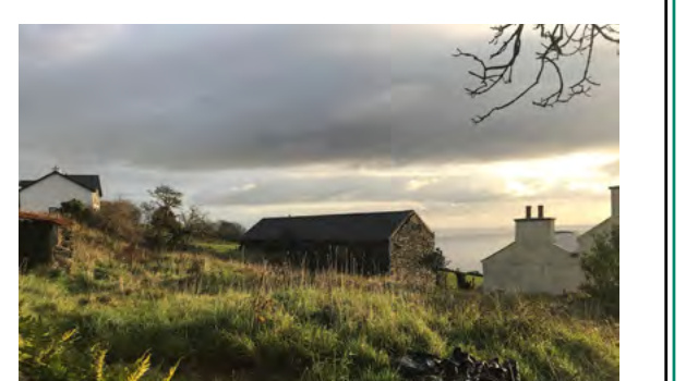 A photograph depicting a rural landscape featuring a large barn-like structure and residential buildings under a cloudy sky.
