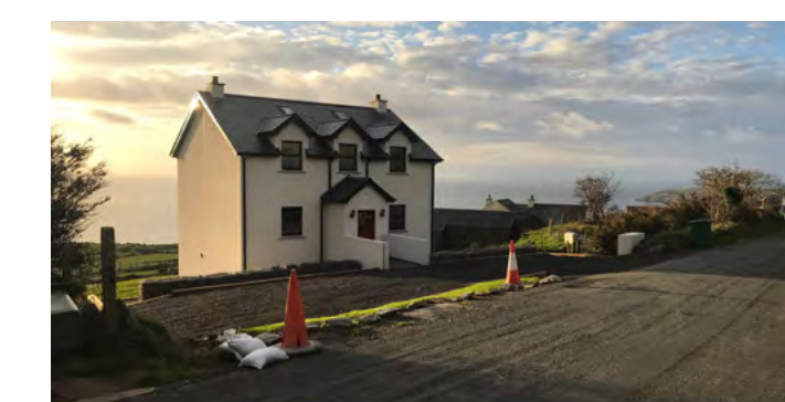 A photograph showing a newly constructed white detached house with dormer windows, situated on a slope with a coastal view in the background.