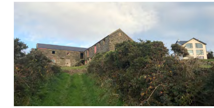 A photograph showing a large stone barn-like structure on a grassy slope with a modern white house visible in the background to the right.