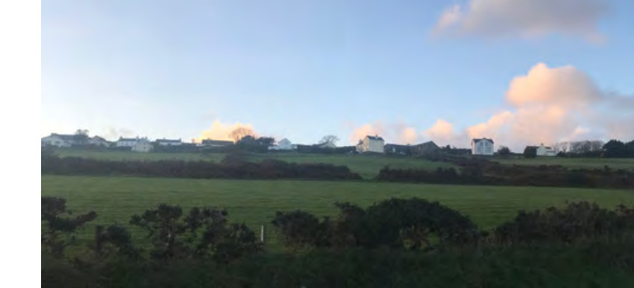 A landscape photograph showing green agricultural fields in the foreground with a row of white residential houses situated on a distant ridge under a blue sky.