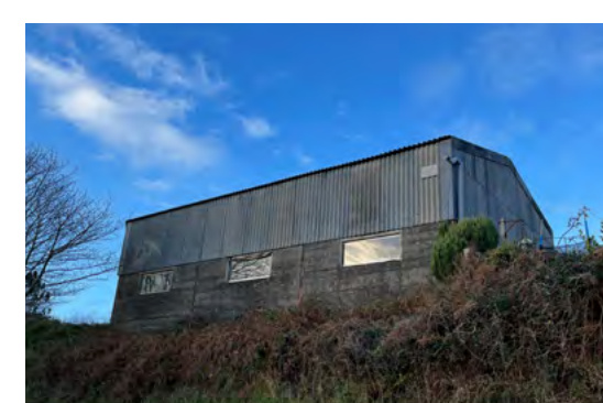 A photograph of a large, weathered agricultural building with corrugated metal siding and a concrete block base, situated on a grassy slope.