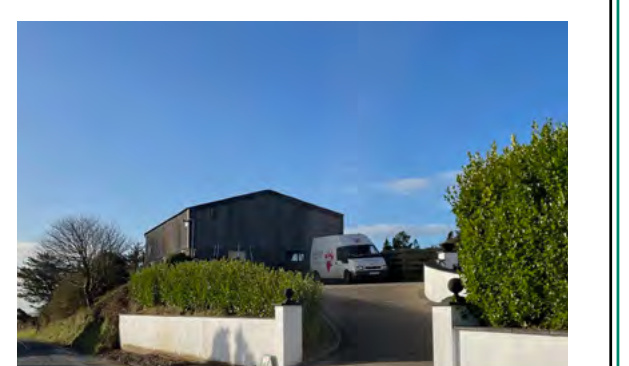 A photograph showing a large dark agricultural-style building on a hill with a white van parked in front, viewed from a driveway with white boundary walls.