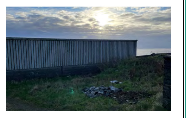 A photograph showing a long corrugated metal shed or outbuilding situated on a grassy slope with a coastal view in the background.