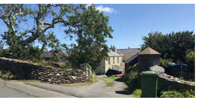 A street-level photograph showing a rural property entrance with a stone wall, driveway, and a two-story house alongside a stone outbuilding with a conical roof.