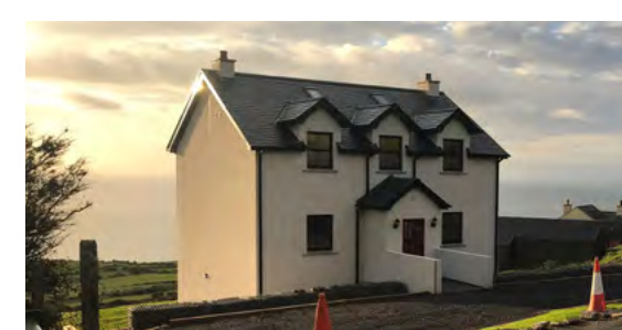 A photograph of a white two-story detached house with dormer windows, situated in a rural coastal location with a view of the sea.