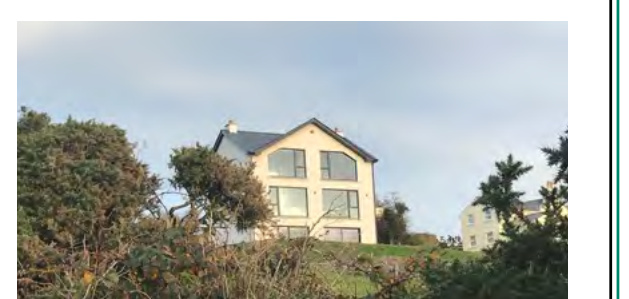 A photograph showing a two-story detached house with large windows situated on a grassy slope, partially obscured by foreground vegetation.