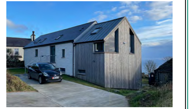 A photograph showing a modern residential building featuring a mix of white render and grey timber cladding with a pitched roof and skylights, with a car parked on the driveway.