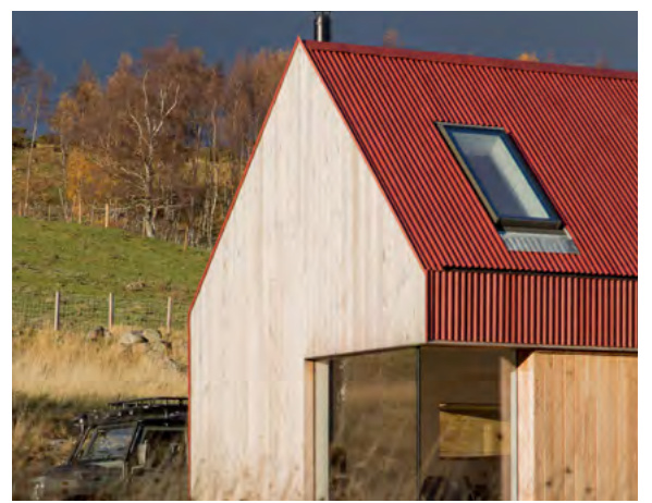 A close-up exterior view of a modern dwelling featuring a red corrugated metal roof, light timber cladding, and a skylight against a rural hillside background.