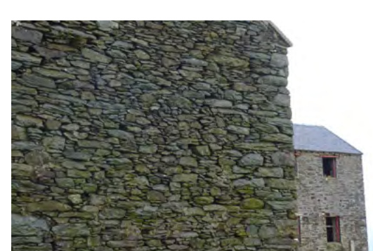 A close-up photograph showing a rough stone wall in the foreground with a stone building featuring window openings visible in the background.
