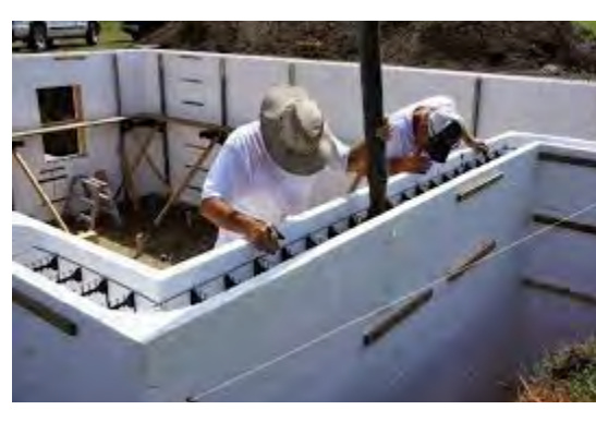 A photograph showing construction workers building white concrete block walls, likely the foundation or lower level of a new structure.
