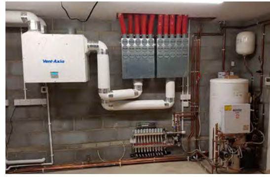 A photograph of a utility room interior showing a Vent-Axia ventilation unit, underfloor heating manifolds with red pipes, and a hot water cylinder.