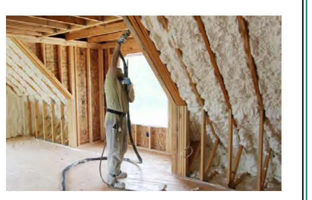 A construction worker is shown spraying foam insulation onto the wooden rafters and walls of a roof space during the building process.