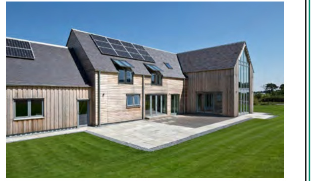 A photograph of a modern, timber-clad residential building featuring a slate roof with solar panels. The property includes a paved patio area and is situated in a rural, grassy setting.