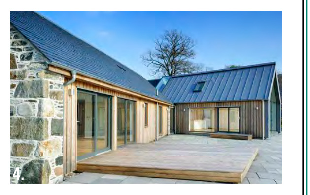 A photograph showing a modern residential extension with wooden cladding and large glass doors attached to an existing stone building, featuring a wooden deck and paved patio area.