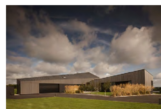 A photograph showing a modern, single-story detached dwelling with dark cladding and a large garage door, set against a cloudy sky with landscaped foreground.
