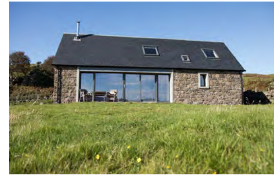 A photograph showing a stone building with a large modern glass extension set in a grassy rural field under a blue sky.