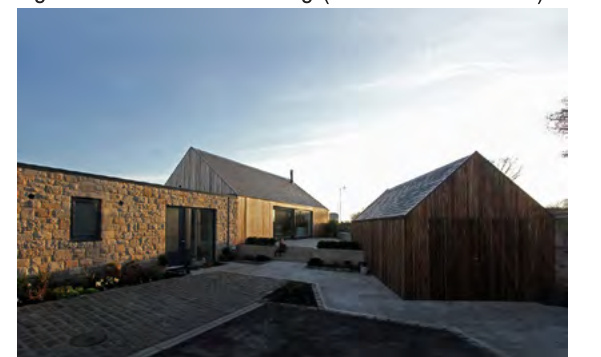 A photograph showing a modern residential property with stone and timber cladding, featuring a paved driveway and an adjacent wooden outbuilding.