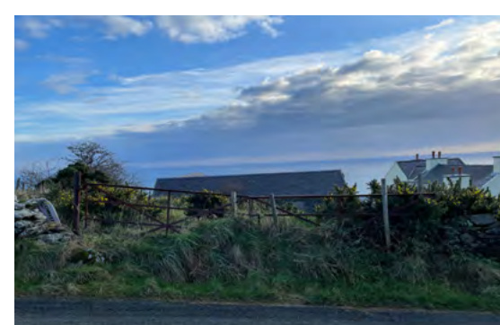 A street-level photograph showing existing rural buildings, including a long barn-like structure and a white house, situated behind a wooden fence and vegetation.