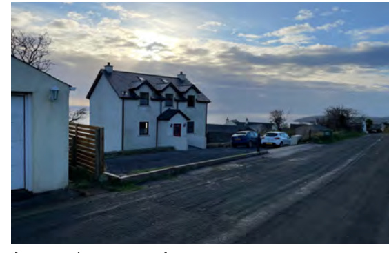 A photograph showing a white two-story detached house with a driveway and parked cars, situated next to a road at dusk.