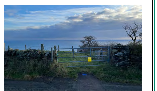 A photograph showing a metal gate and stone wall leading to a coastal field with the sea in the background.