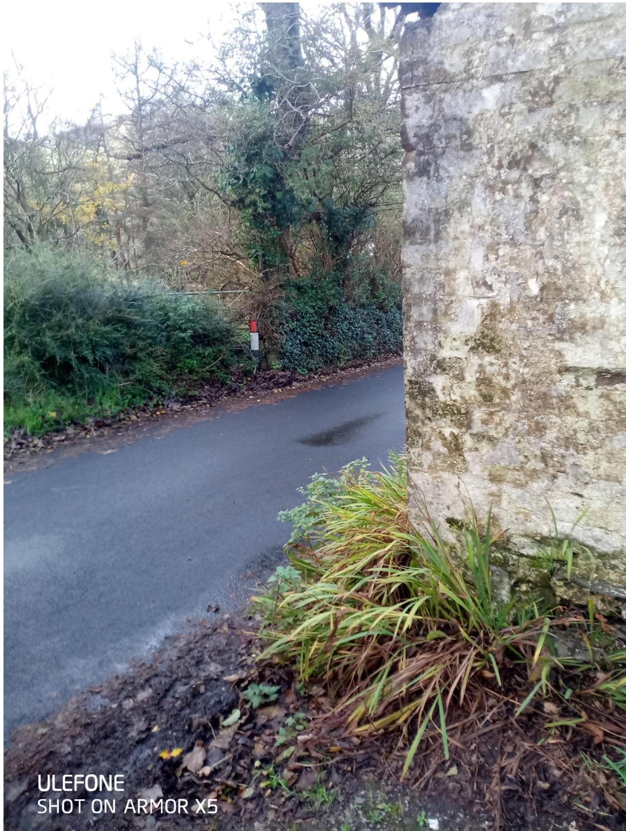 A photograph showing a rural road curving to the left with a stone wall on the right and vegetation on the left.