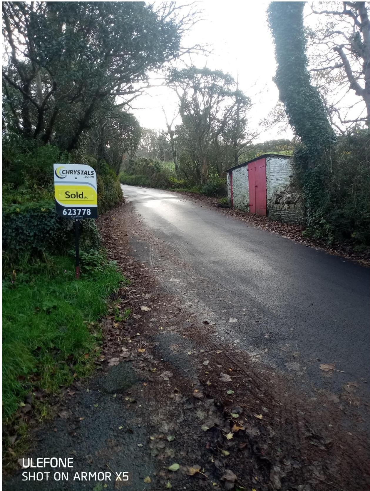 A photograph of a rural road showing a stone outbuilding with red doors on the right and a 'Sold' estate agent sign on the left.