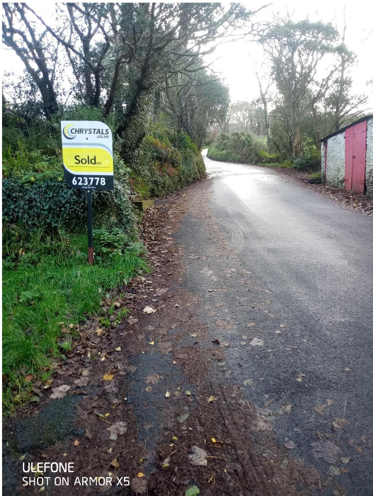 A photograph showing a rural road with a 'Sold' sign on the left and a small outbuilding with red doors on the right.