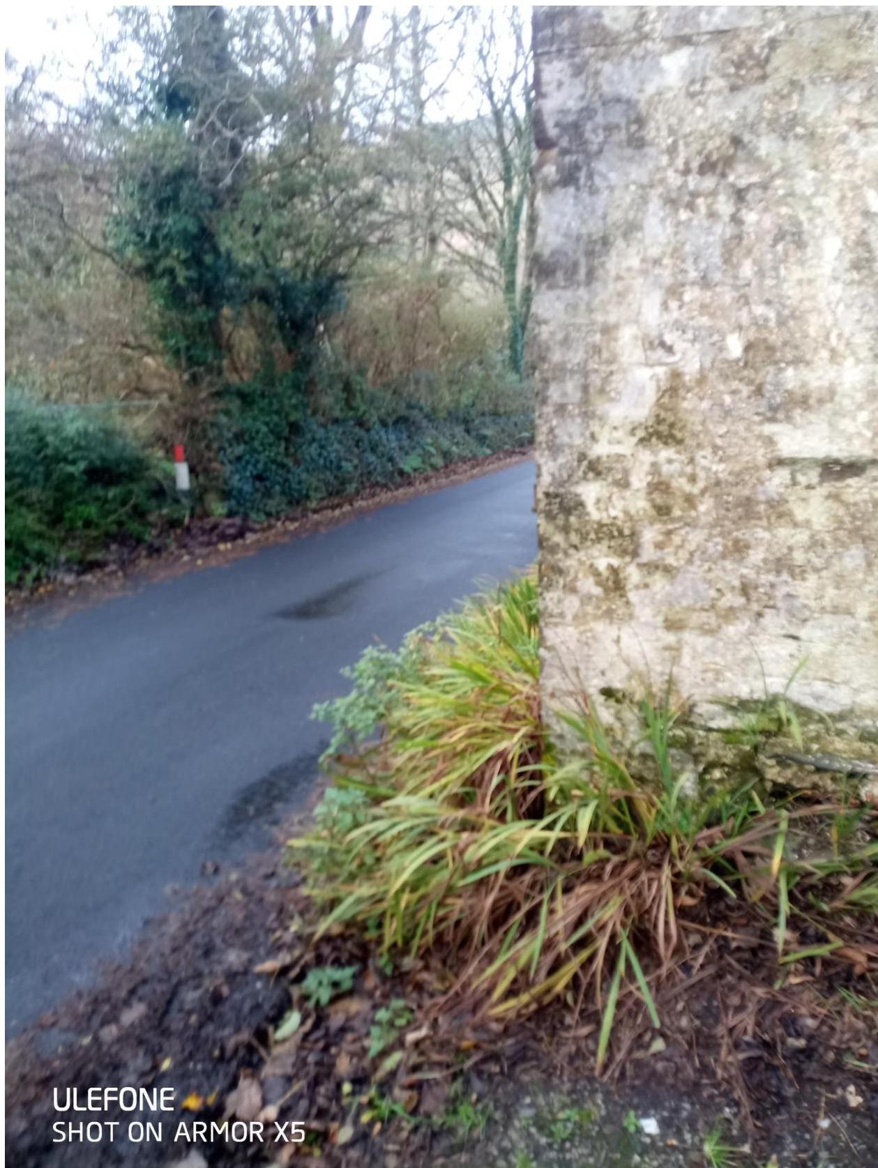 A photograph showing a paved road curving past a rough stone wall on the right, with trees and vegetation in the background.