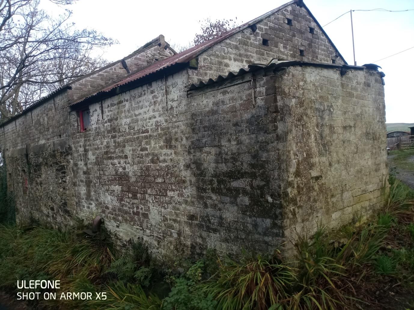 A photograph of a weathered stone outbuilding with a corrugated roof, showing signs of disrepair and overgrown vegetation.