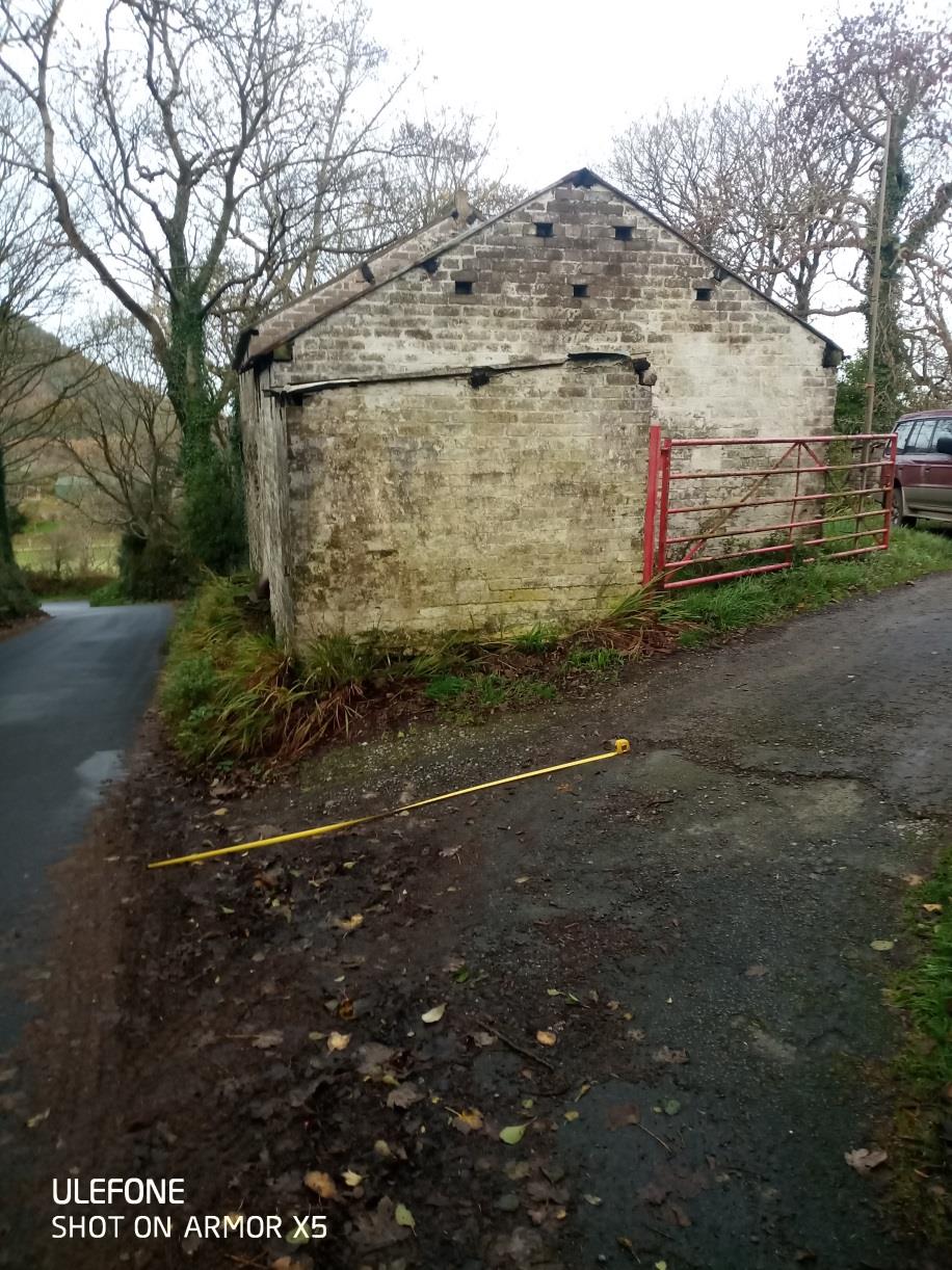 A photograph showing a weathered stone outbuilding situated next to a rural road, featuring a red metal gate and a yellow measuring tape on the ground.