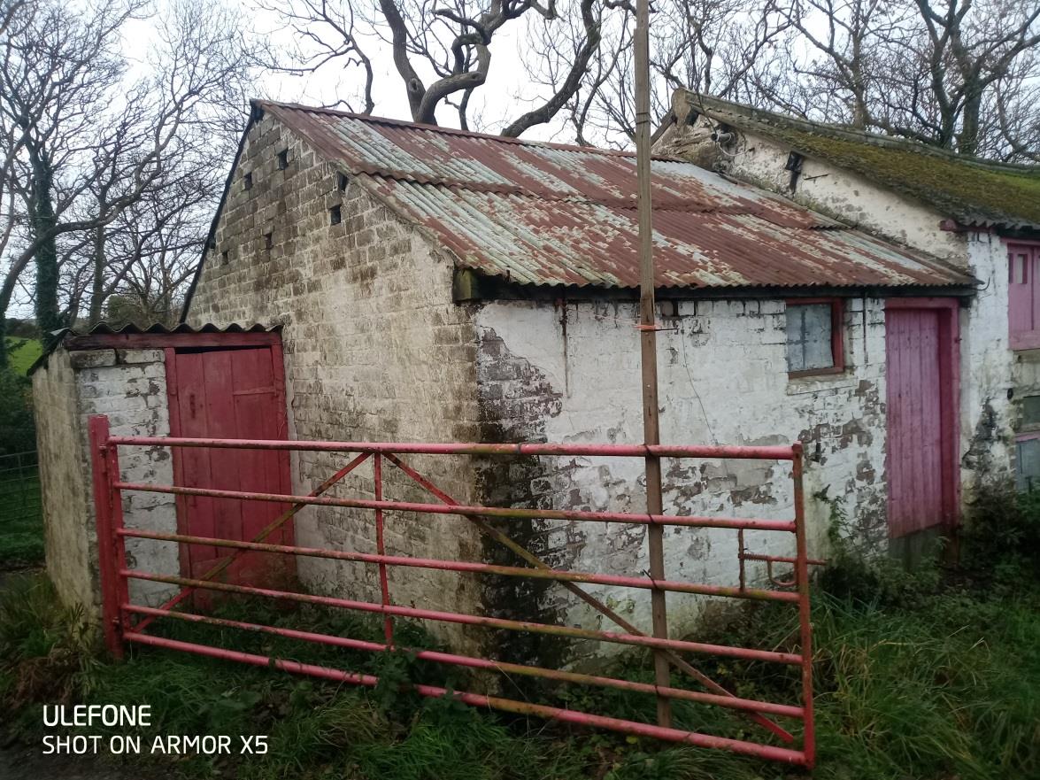 A photograph showing a dilapidated stone outbuilding with a rusty corrugated roof and red doors, situated behind a red metal gate in a rural setting.
