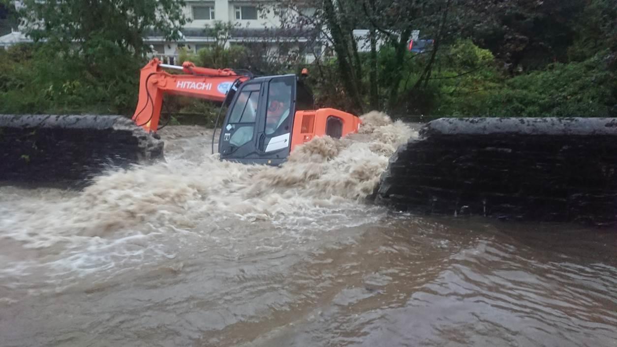 A photograph showing an orange excavator partially submerged in rushing floodwater between stone embankments.