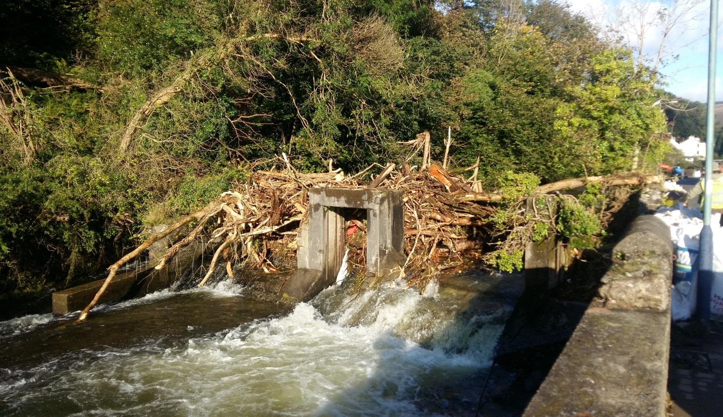 A photograph showing a river channel with a concrete outlet structure heavily obstructed by flood debris including logs and branches.
