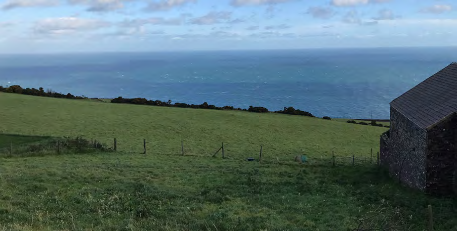 A photograph showing a grassy field sloping down towards the sea with a stone building visible on the right edge.