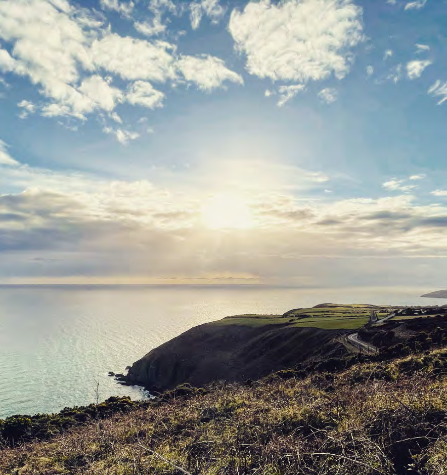 A scenic photograph of a coastal landscape featuring a grassy cliff edge overlooking the sea with a road winding along a distant headland under a bright sky.