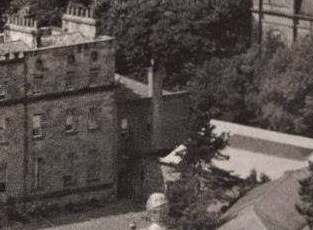 A black and white photograph showing the exterior of a large, multi-story stone building, likely Castle Mona, surrounded by trees and a wall.