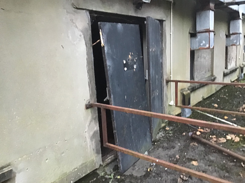 A close-up photograph of a building's exterior wall showing a dark doorway and adjacent windows that appear boarded up or covered, with a rusty metal railing in the foreground.