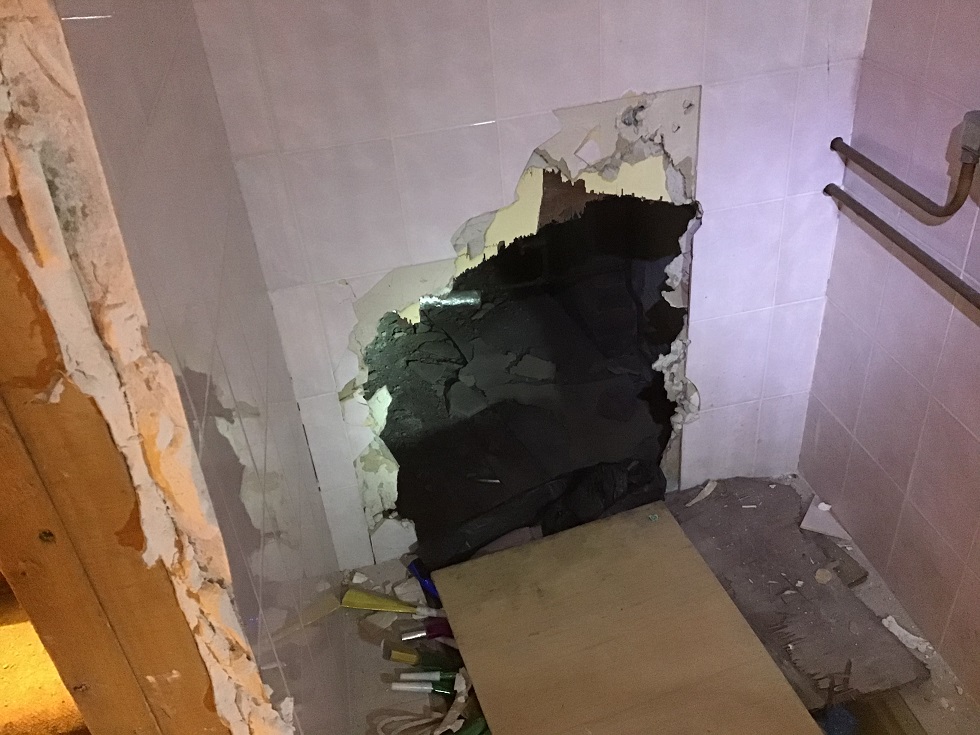 A photograph showing a damaged interior wall with white tiles and a large hole revealing dark backing material, surrounded by debris and exposed wooden framing.