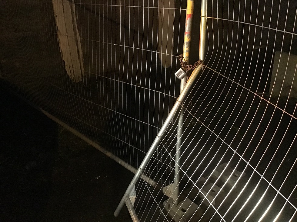A low-light photograph showing a temporary metal mesh security fence secured with a padlock, likely enclosing a construction or demolition site.
