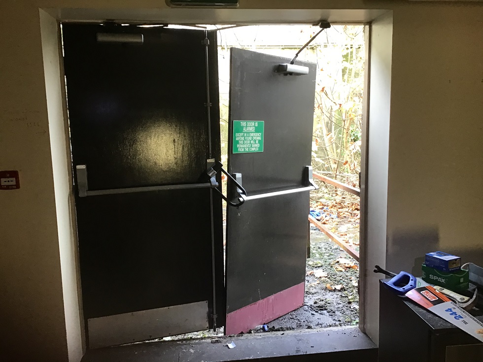 A photograph showing an interior view of a heavy double door with one leaf open, revealing an outdoor area with debris and vegetation, likely during demolition work.