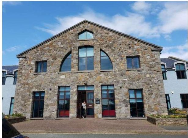 A photograph showing the front elevation of a large stone building with a gable roof and prominent arched windows. A person is standing in the central entrance on a paved forecourt.