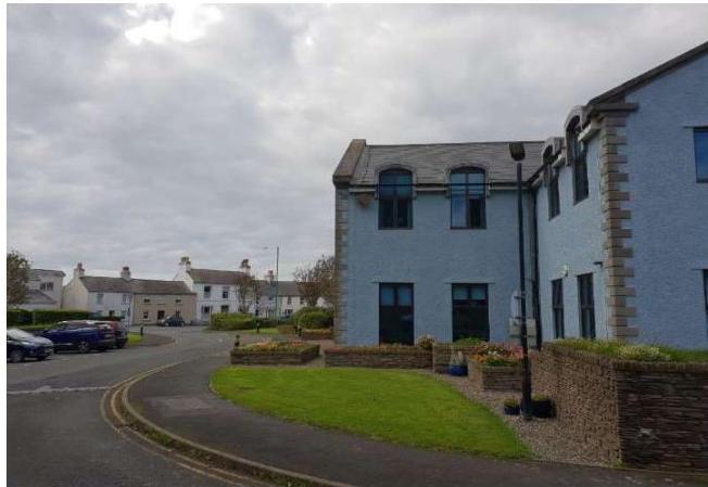 A street-level photograph showing a modern blue detached house with stone quoins on the right, set against a backdrop of traditional white houses and a cloudy sky.