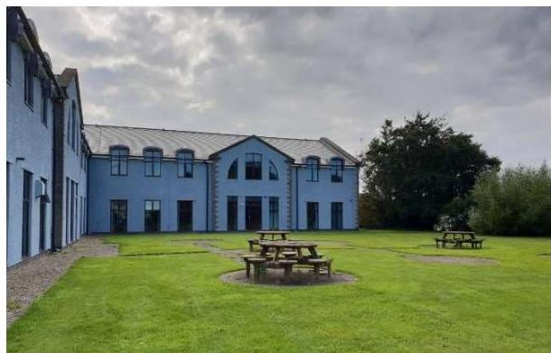 A photograph showing the exterior of a large, light blue building with a grey roof and dormer windows, situated behind a grassy area with wooden picnic tables.