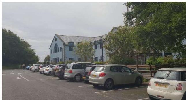 A photograph showing a parking lot with several cars parked in front of a large, light blue building surrounded by trees.